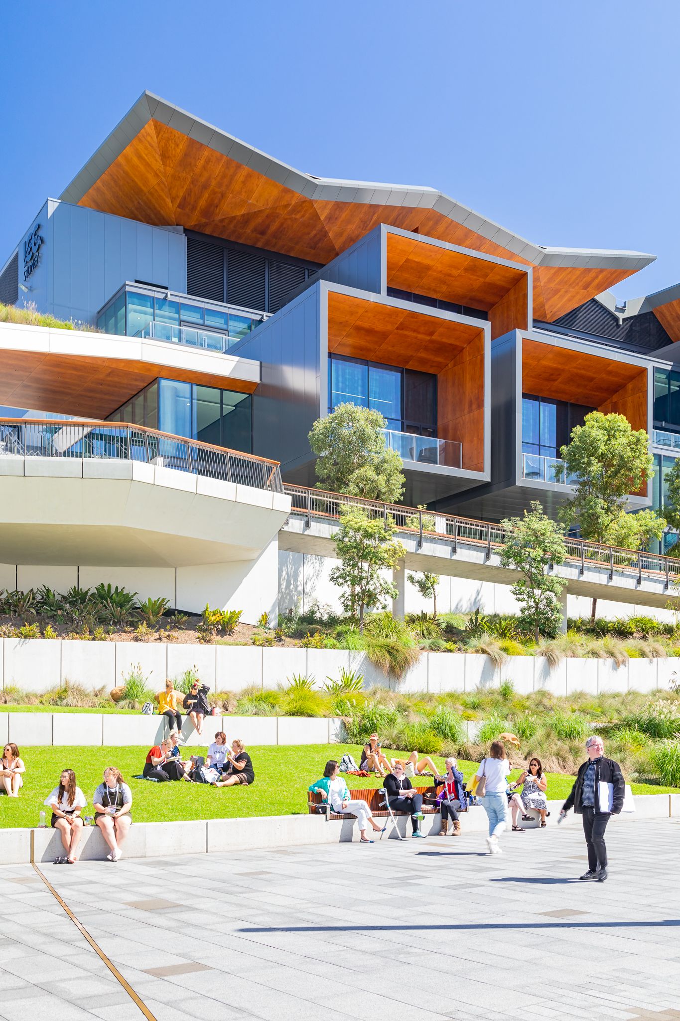 People relax on lawns and steps in front of a contemporary building with angular timber-clad balconies and landscaped terraces.