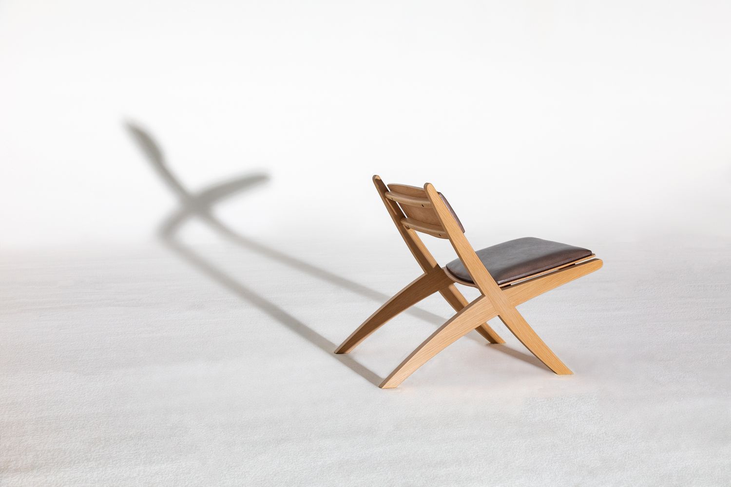 Modern folding wooden lounge chair with a grey cushion casting a long, dramatic shadow on a soft white floor.