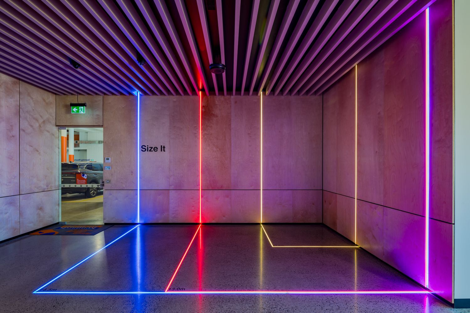 Corridor with a slatted timber ceiling and colorful red, blue and purple LED light lines on the floor and walls beside a Size It sign.