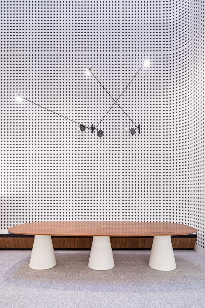 Oval timber table on three white conical bases beneath a modern pendant light on a perforated white wall.