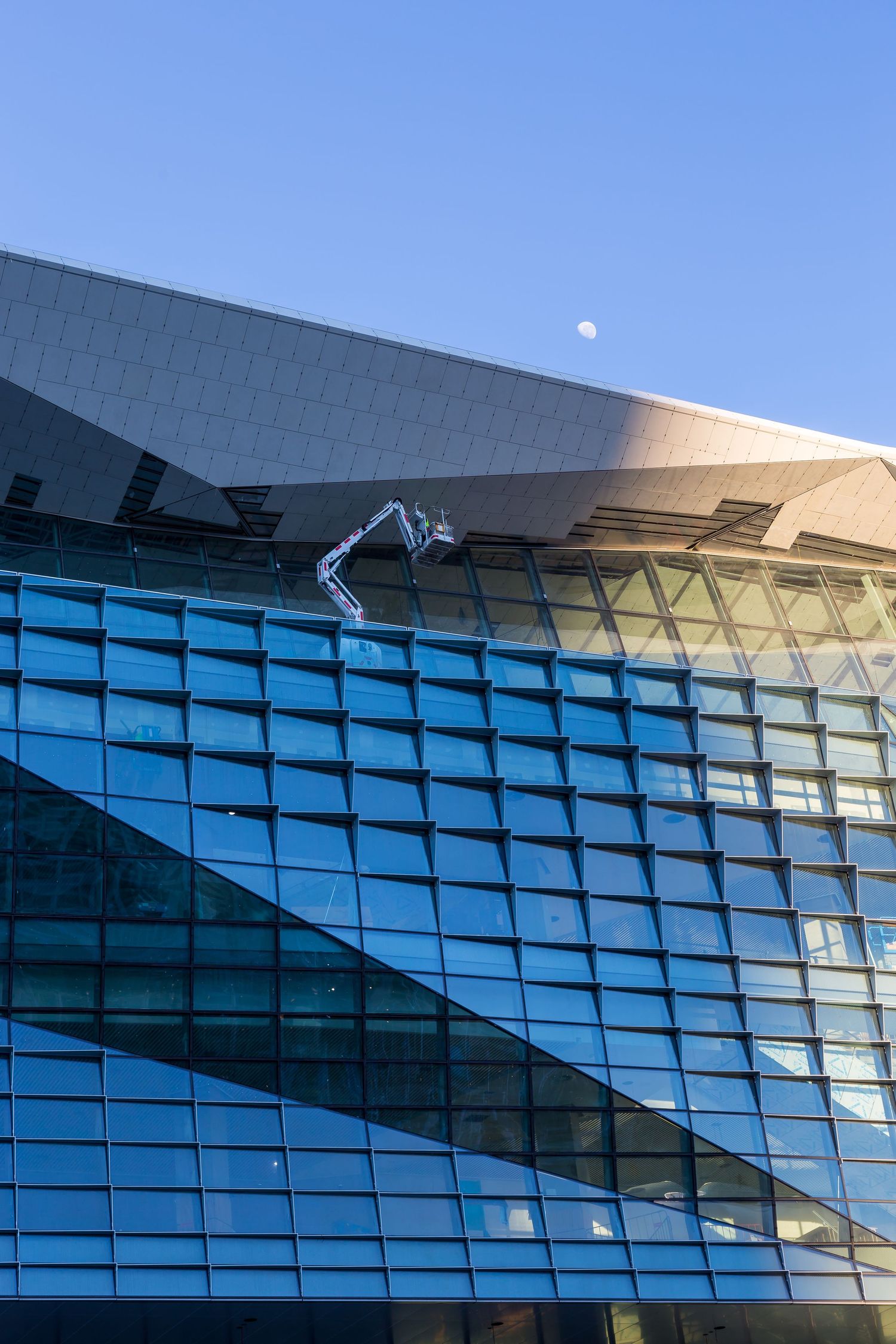 Glass curtain wall building with an angular metal roof, a cherry picker working on the facade and a pale moon above.