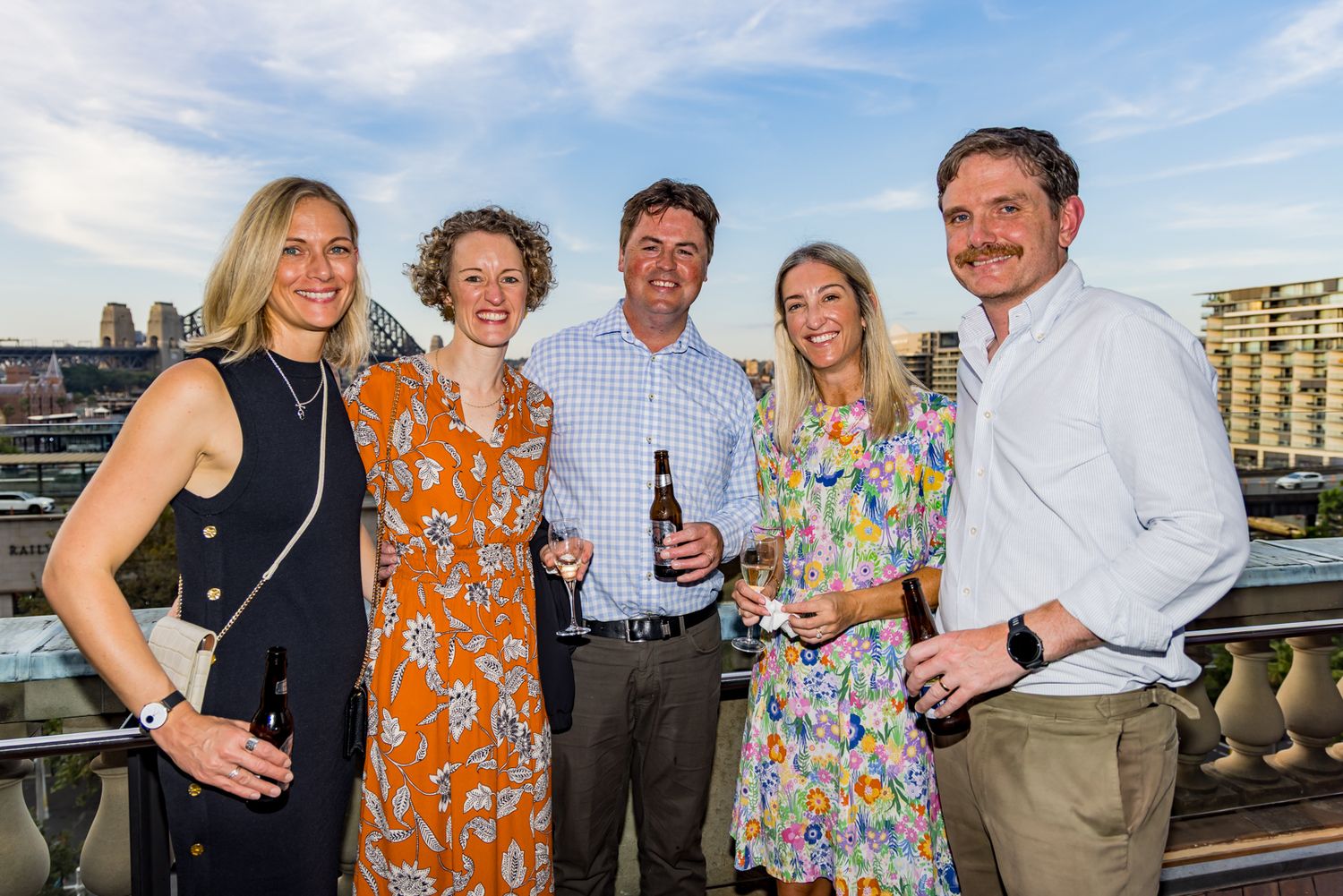 Group of five friends holding drinks and smiling together on a rooftop terrace with a city skyline in the background.
