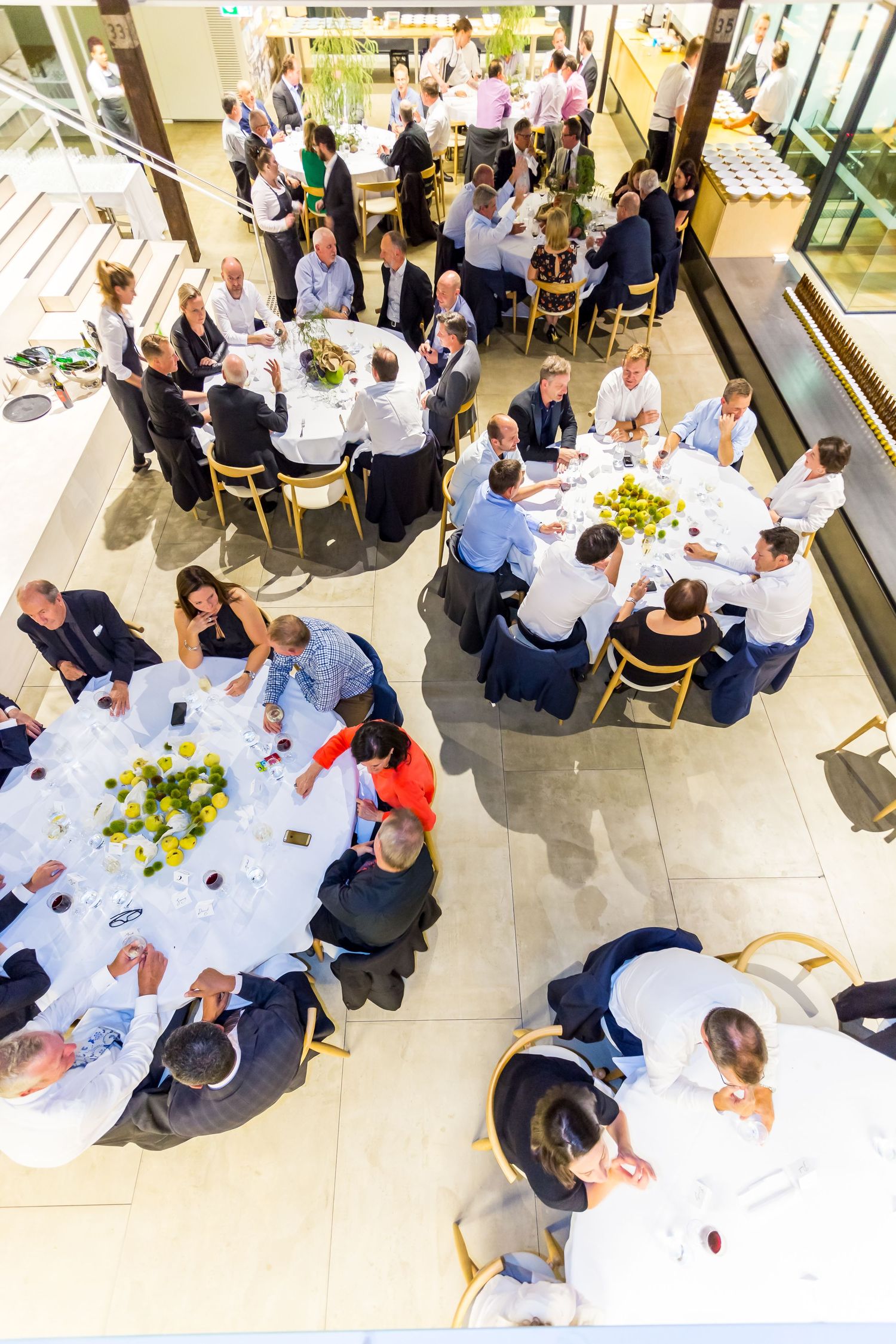 Overhead view of guests seated at round tables with yellow flower centerpieces during a large corporate lunch in a bright, modern venue.