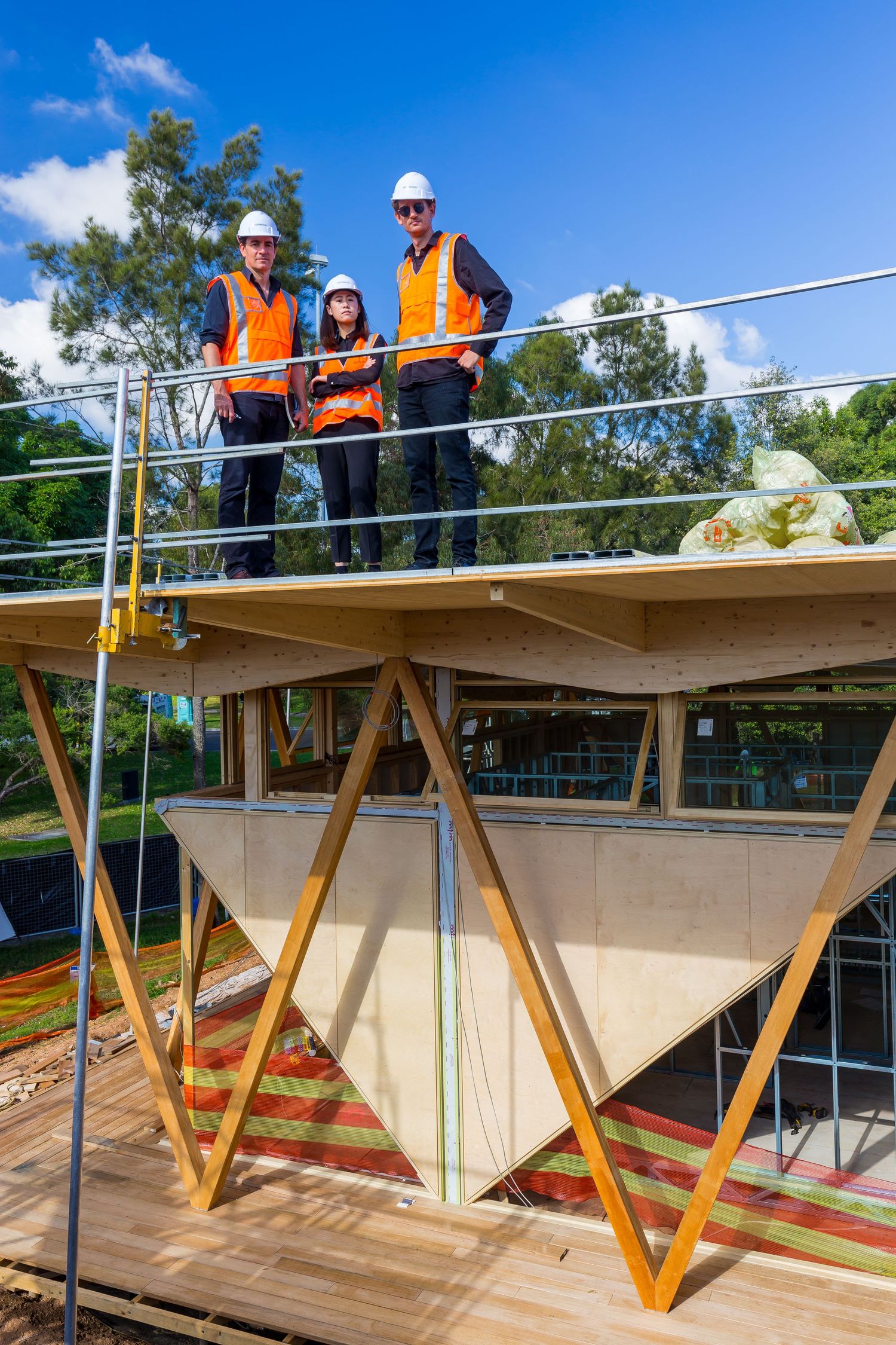 Three people in hard hats and orange high-visibility vests stand on a timber-framed building under construction beneath blue sky.