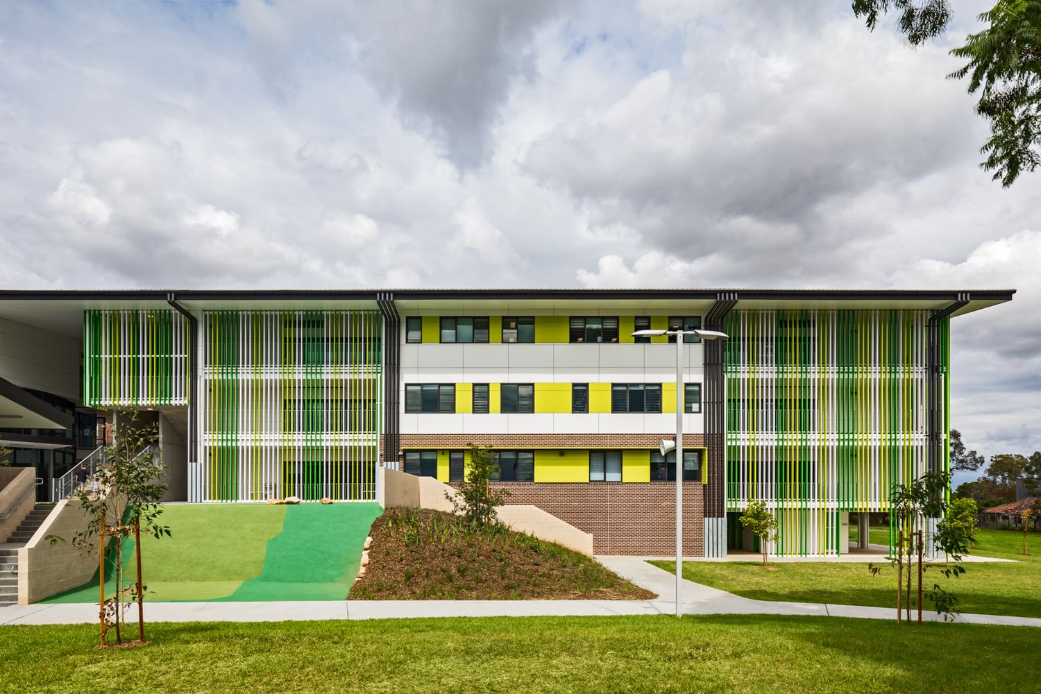 Modern school building with colorful green and yellow facade panels, landscaped grassy slopes and young trees under a cloudy sky.