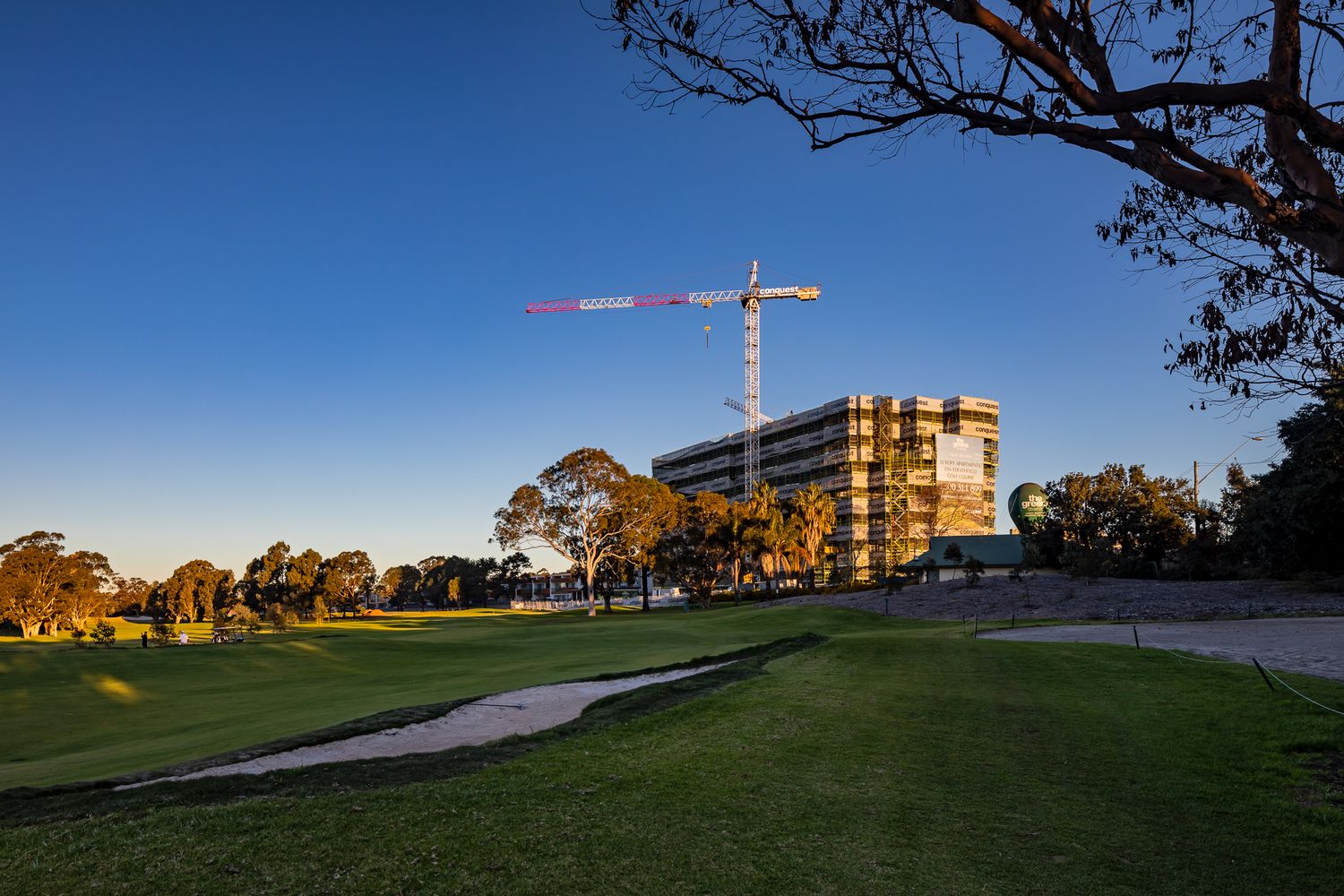 Tall crane beside a multistorey building under construction, viewed across a golf course at sunset with clear blue sky.