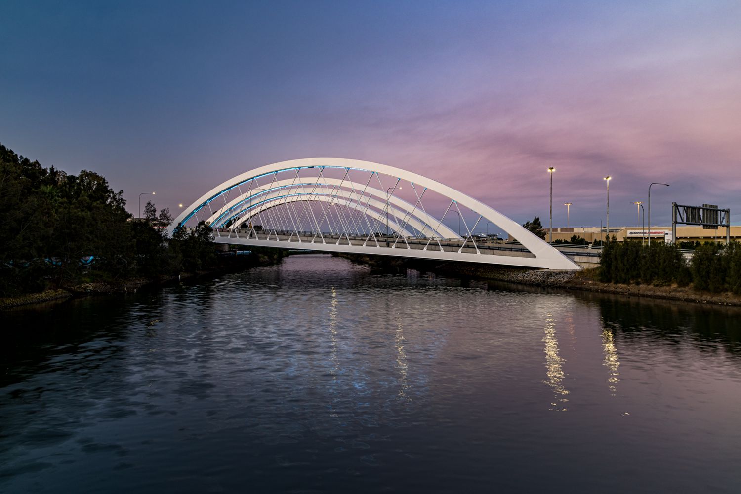 White steel arch bridge spanning a calm river at dusk, with soft pink and purple sky colors reflected in the still water.
