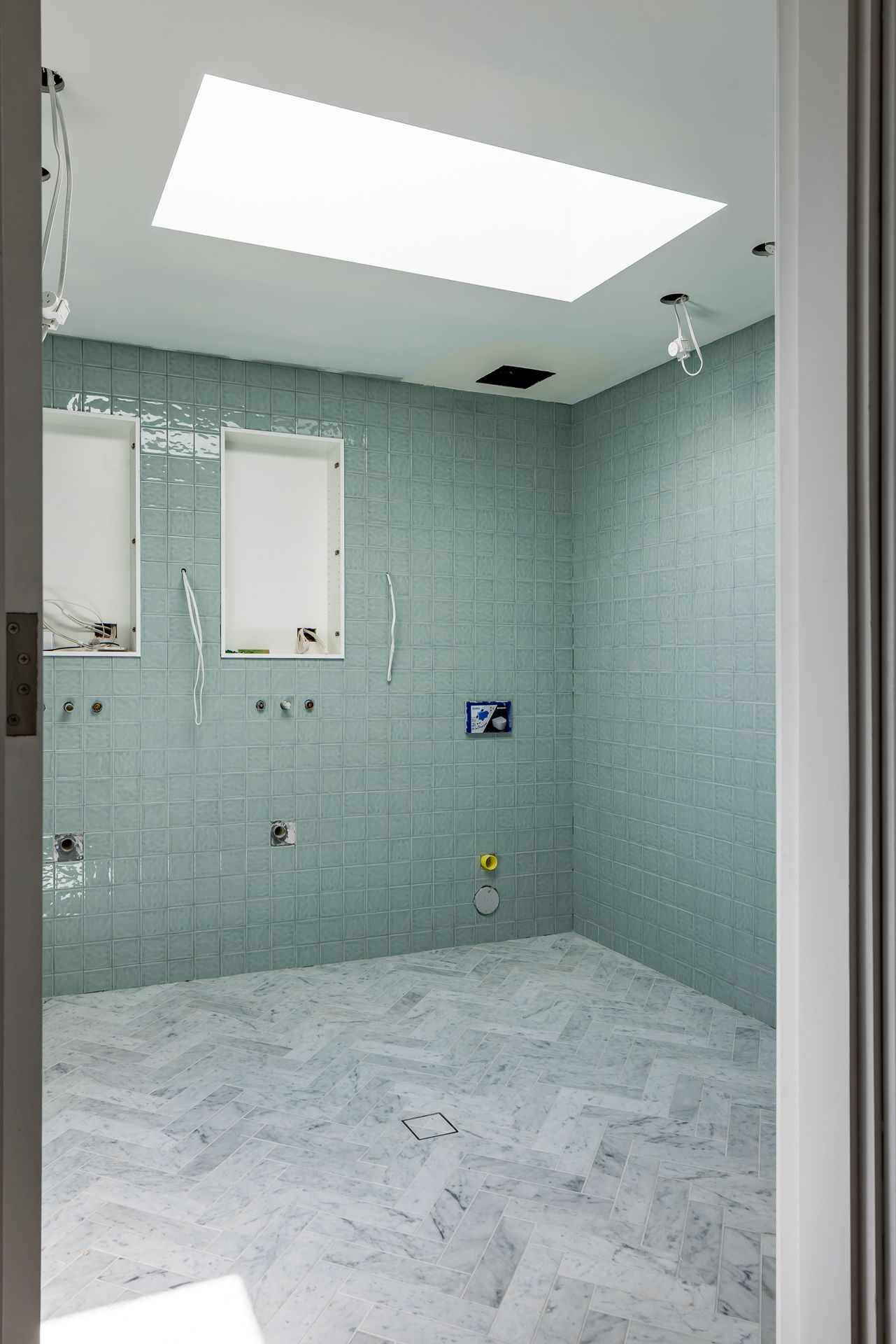 Bathroom under construction with green glass wall tiles, herringbone marble flooring, a large skylight and exposed plumbing fittings.