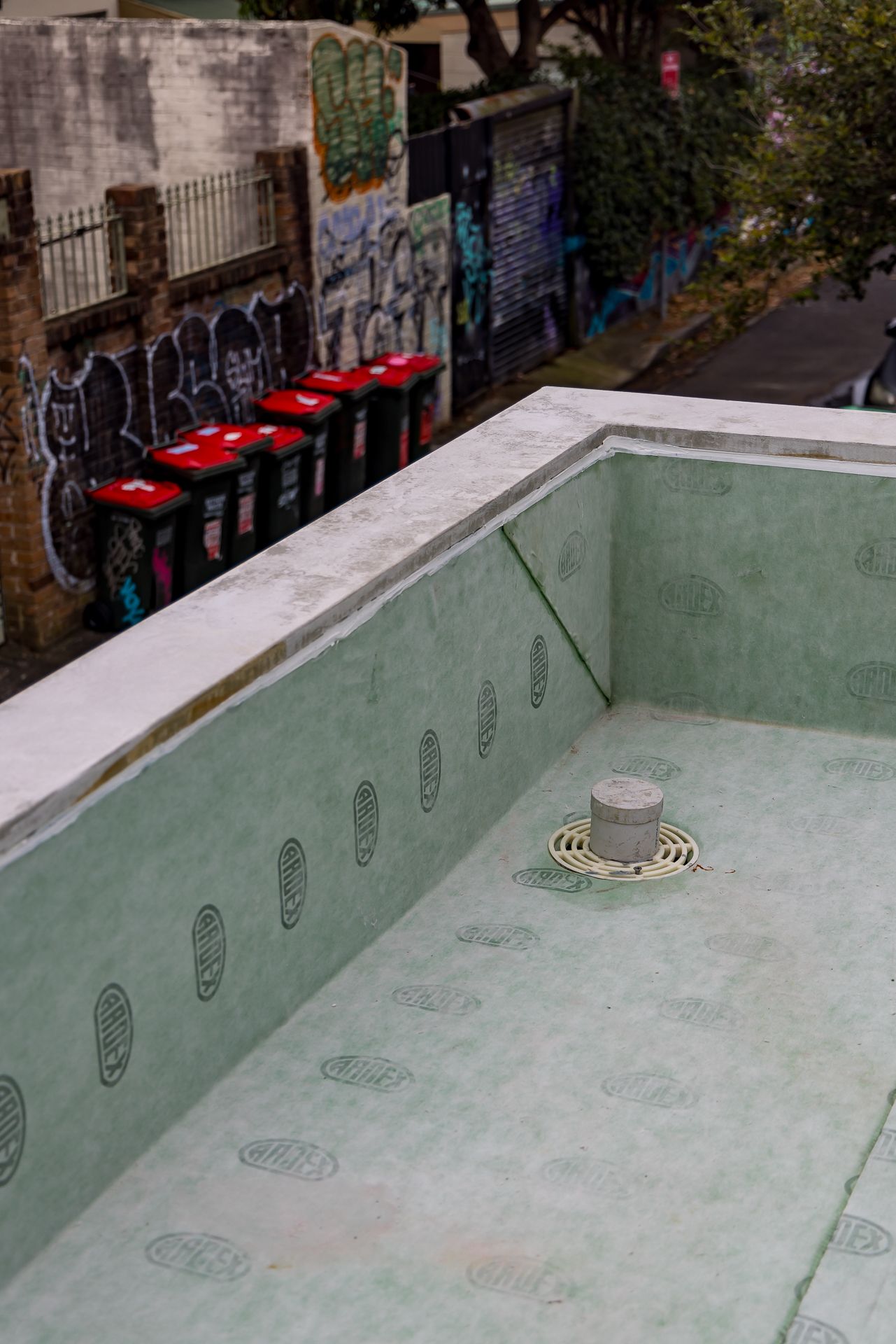 Empty waterproofed rooftop pool shell with green membrane and floor drain overlooking red-lidded bins and graffiti-covered walls.
