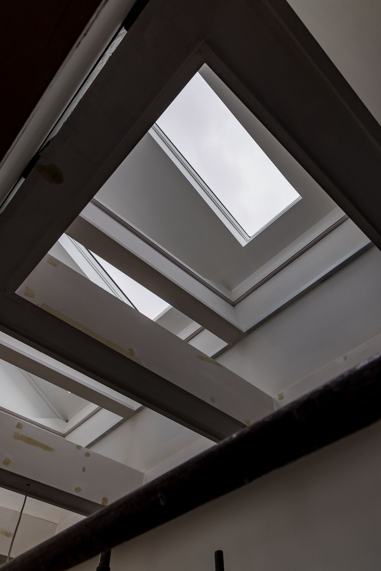 Looking up through angled skylight wells and white structural beams toward a bright overcast sky.