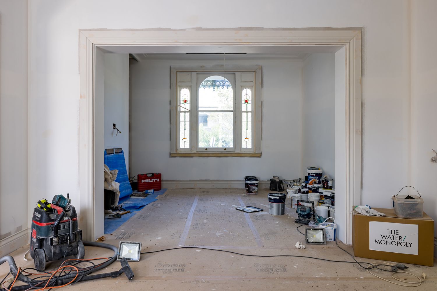 Renovation in progress in a spacious living room with exposed subfloor, power tools and equipment, and an arched window in the background.