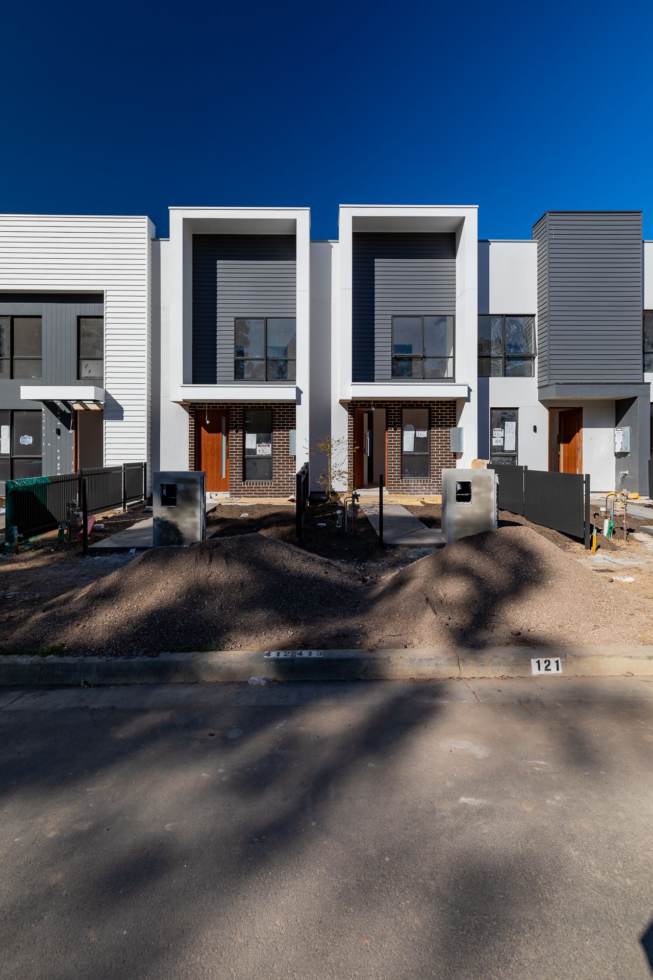 Modern two-storey townhouses with white and gray facades, orange front doors and fresh soil mounds along a newly built street.
