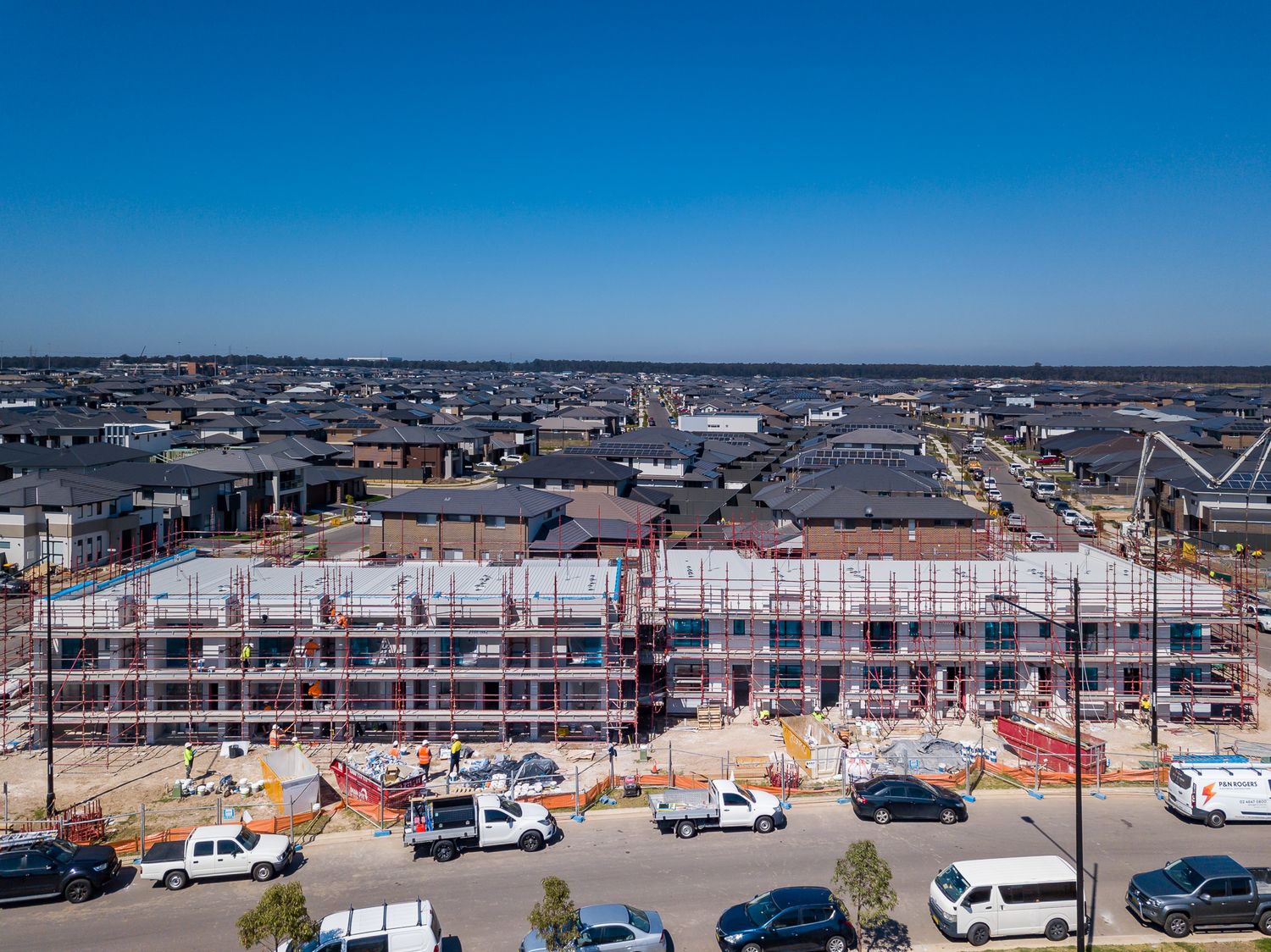 Aerial view of townhouses under construction wrapped in scaffolding with work vehicles parked in front and a suburban neighbourhood beyond.