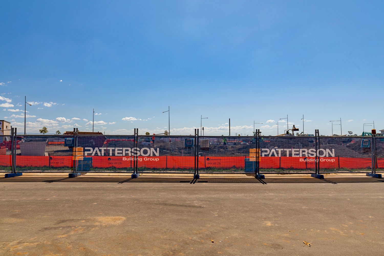 Blue sky above a Patterson Building Group construction fence with red and blue hoarding panels along a dirt road.