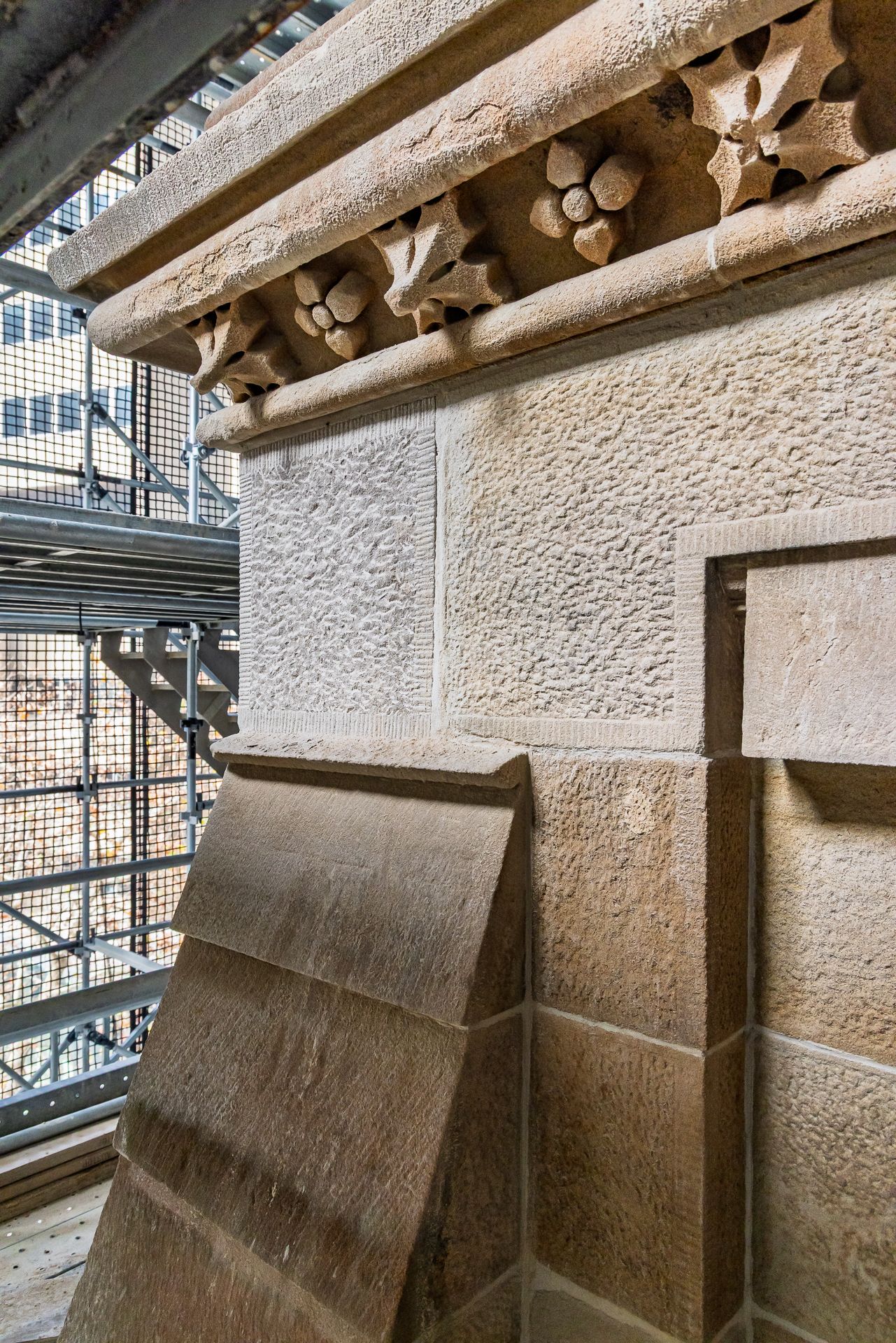 Restored stone cornice with carved four-petal flower ornaments above textured sandstone walls inside a scaffolded structure.