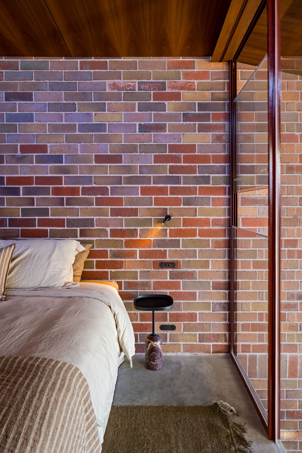 Bedroom detail showing a multicolored brick wall, white linen bedding, a black side table, a wall reading light, and a woven rug.