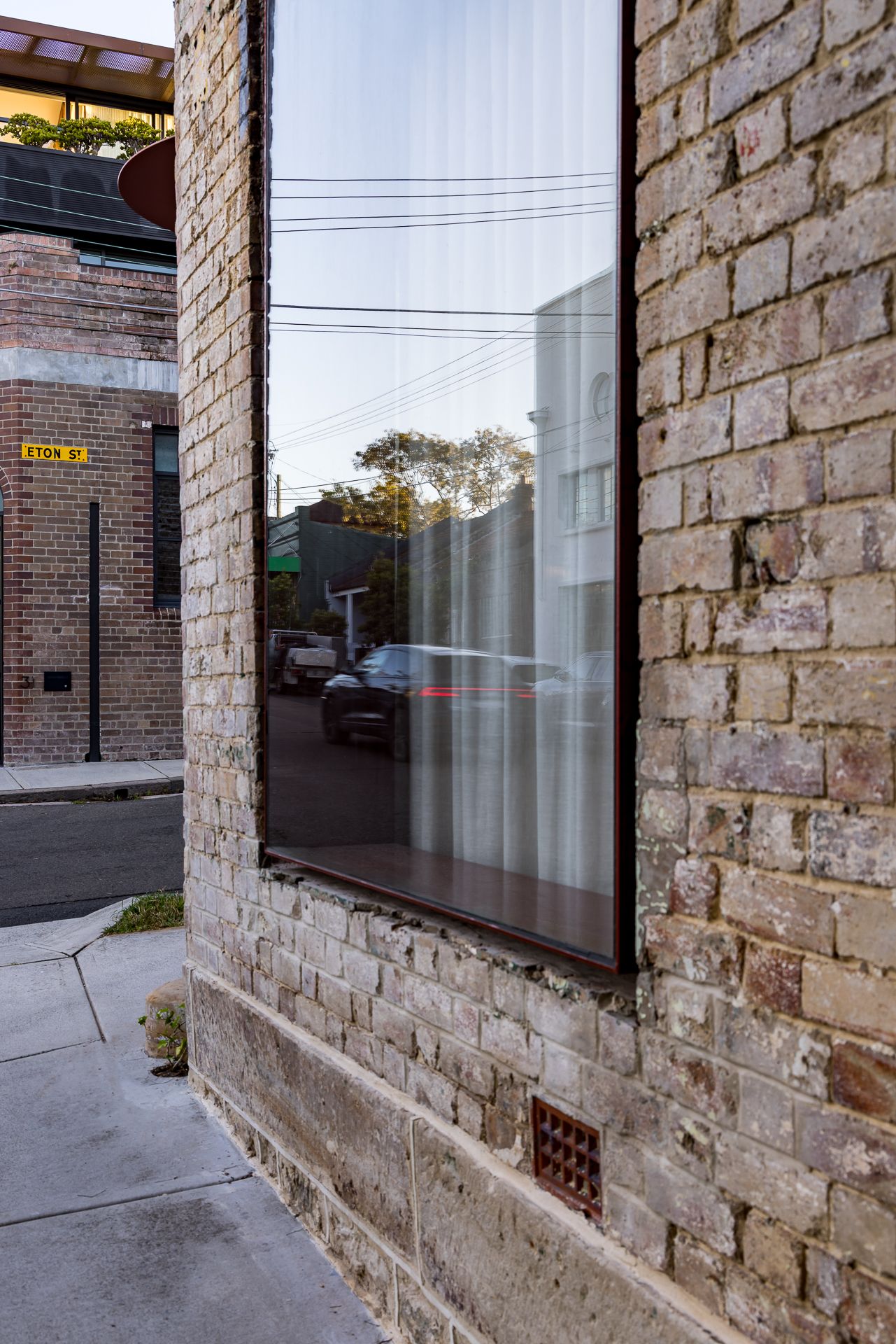 Corner of an old brick building with a large dark-framed window reflecting a quiet street, parked cars and power lines.