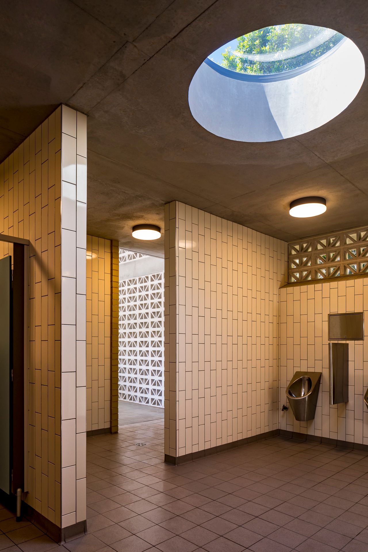 Interior of a public restroom with cream vertical tiles, a circular skylight, breeze block screens and wall-mounted urinals.