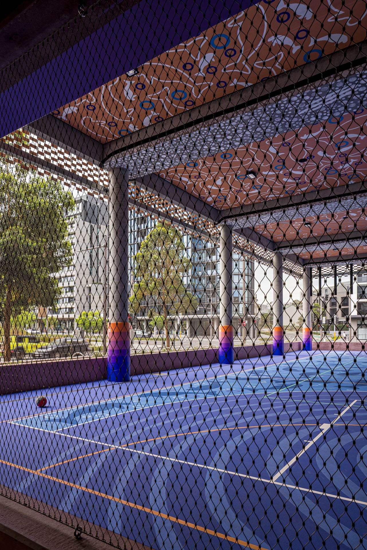 Covered outdoor basketball court with blue surface, colorful painted columns, patterned ceiling and mesh fencing beside urban apartment buil