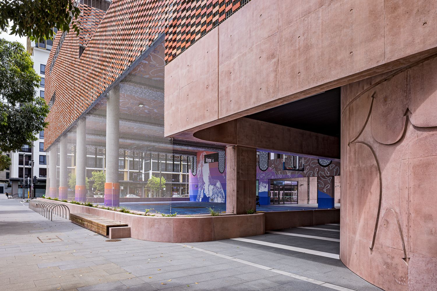 Pink concrete building facade with etched artwork beside a colorful covered court, paved walkway and slender columns.