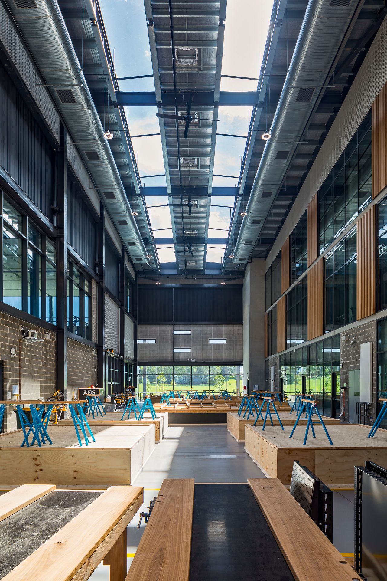 Tall light-filled atrium with skylights, timber workbenches, blue stools and glazed walls leading to a green outlook.