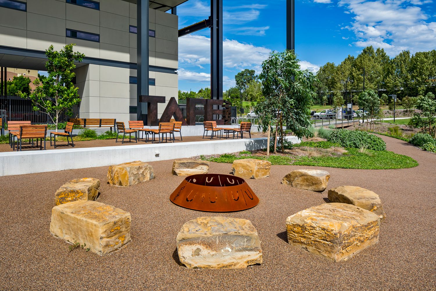 Campus courtyard with large sandstone boulders circling a steel fire pit, with benches, young trees and a modern building behind.