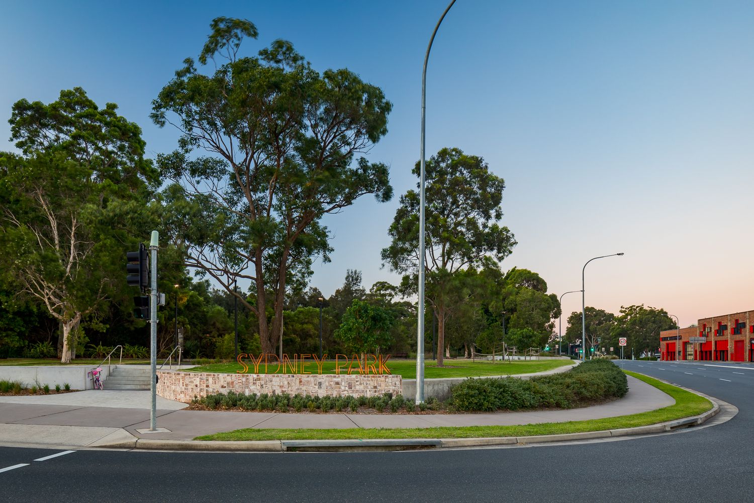Sydney Park entrance corner at dusk, with a sandstone feature wall, hedges, tall gum trees and a curving road under a pastel sky.