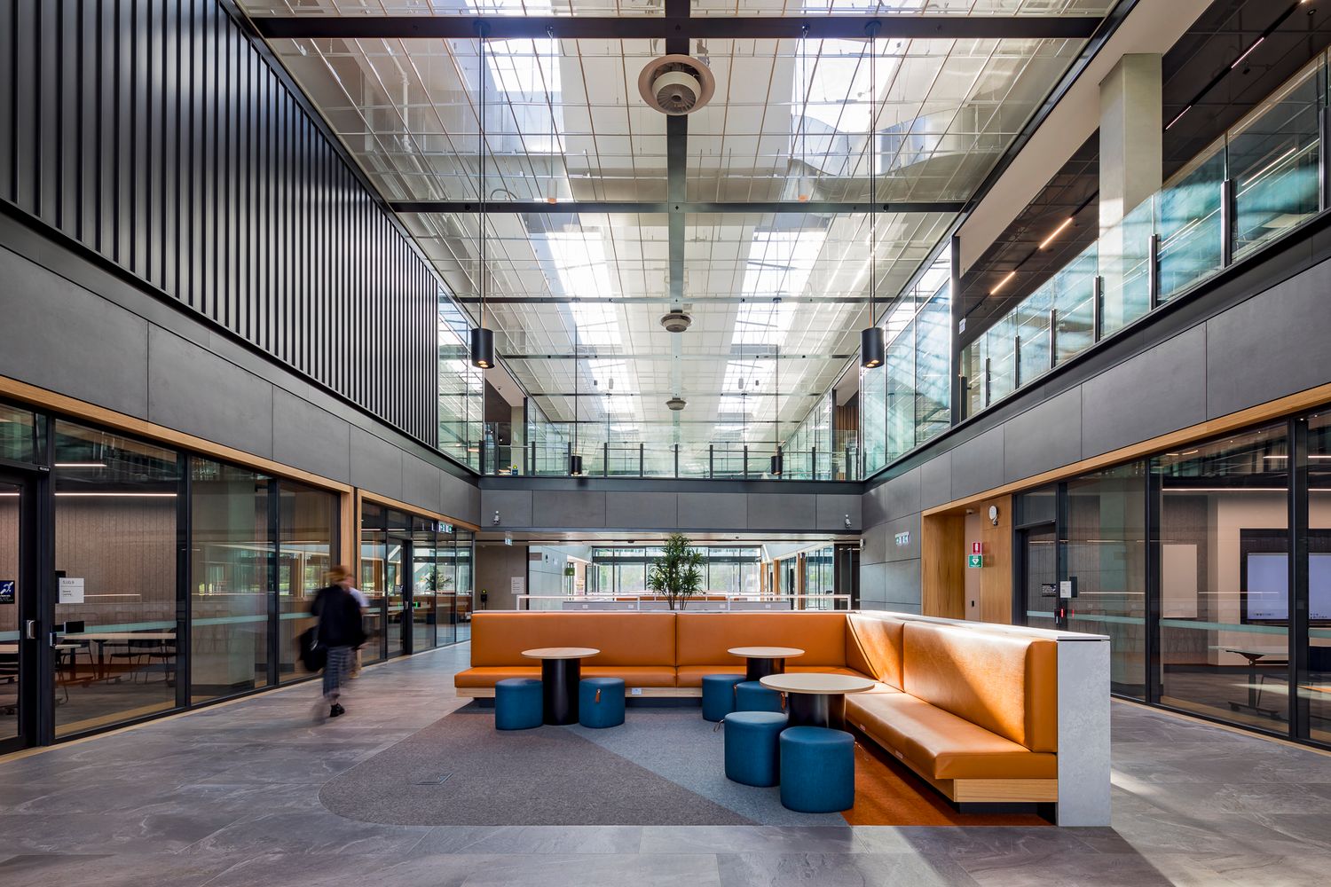 Bright multi-level atrium at a TAFE campus with orange bench seating, blue stools, skylights and students walking past glass-fronted rooms.