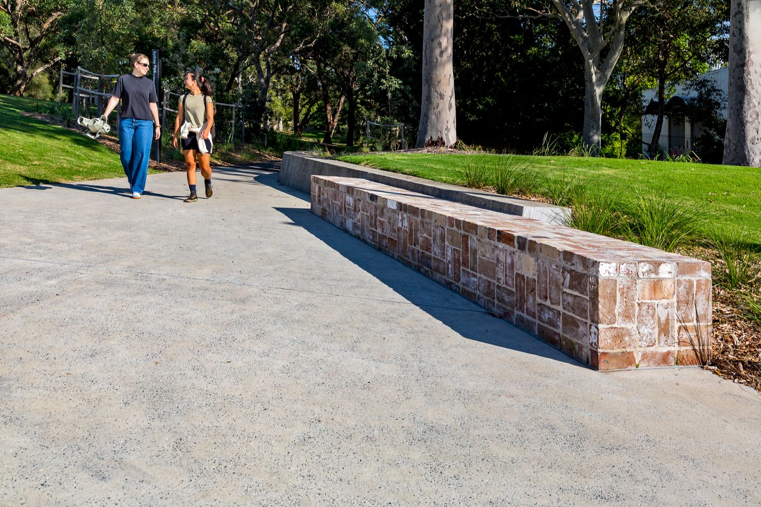 Two people walk along a wide concrete path beside a long sandstone block seating wall in a landscaped park.