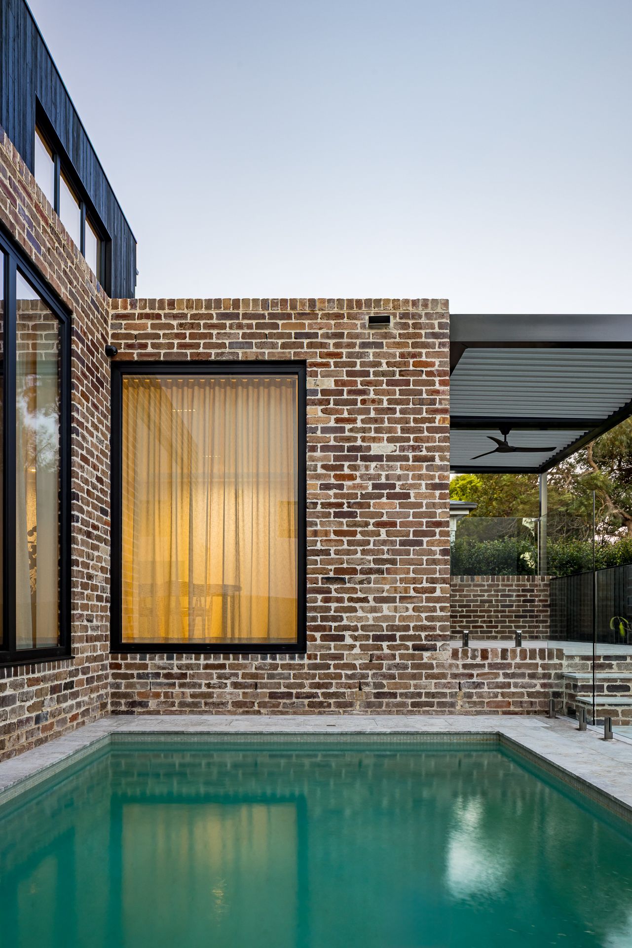 Modern brick home with black-framed windows and a warm glowing curtain beside a turquoise swimming pool and pergola.