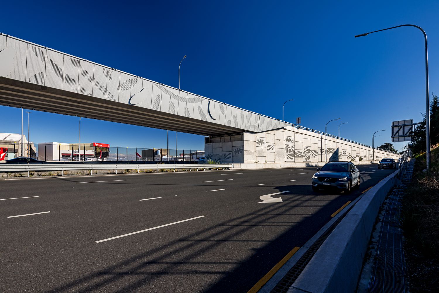A car drives along a wide road beneath a modern elevated bridge with patterned perforated panels and long shadows on a sunny day.
