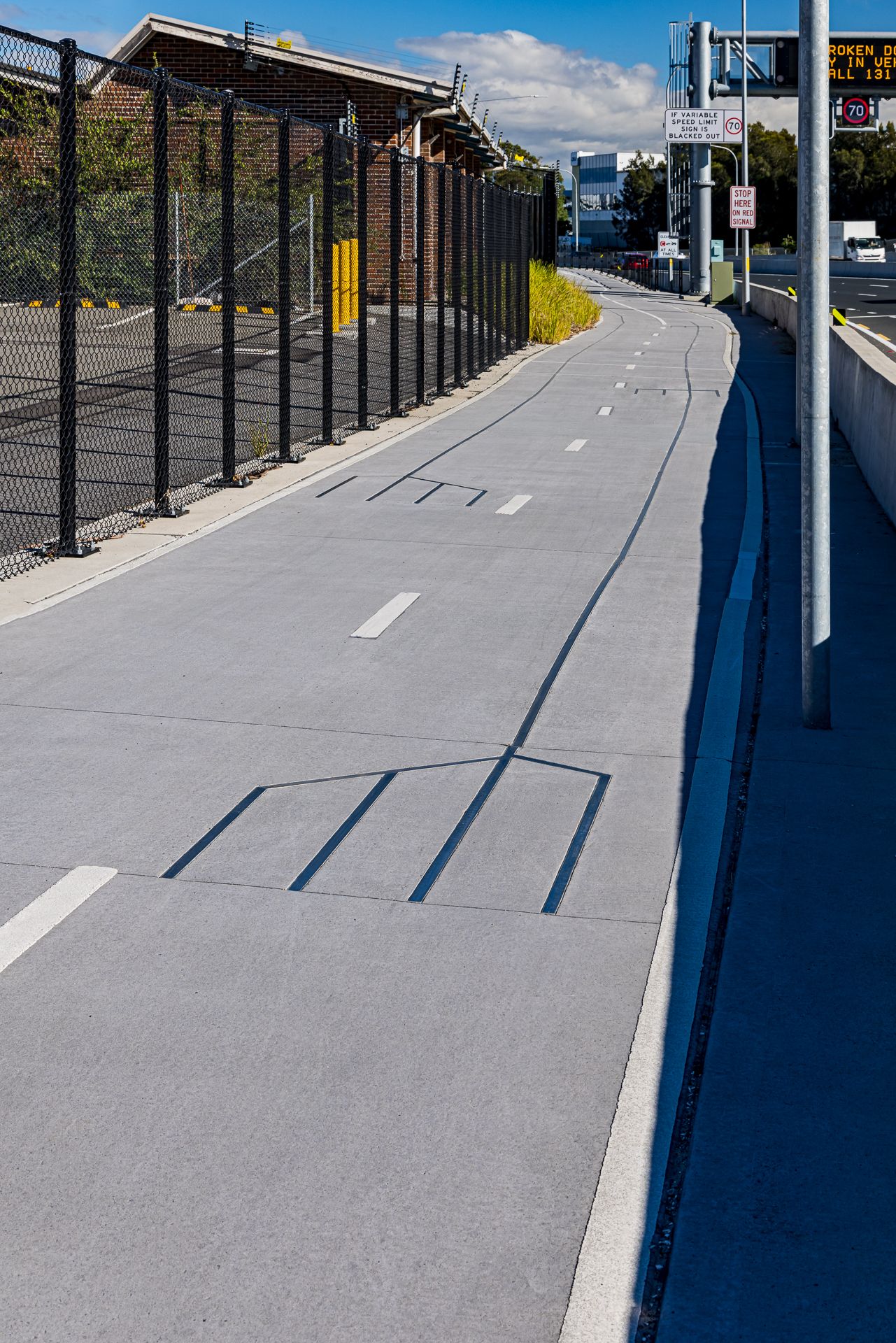 Smooth grey shared cycle path with painted lane markings running alongside a black security fence and a motorway with overhead signs.