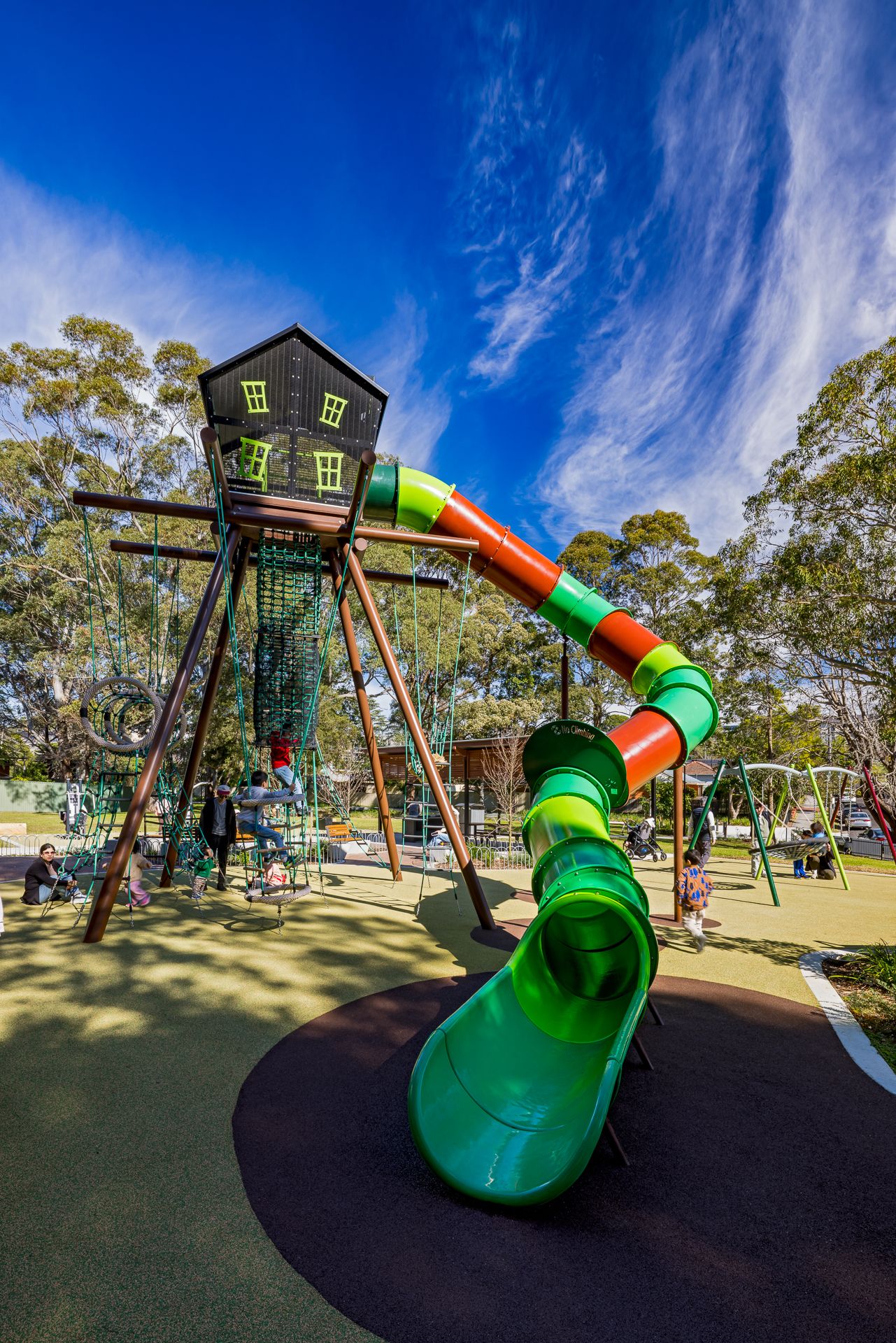 Colorful playground with a treehouse tower, tall green and orange tube slide, and climbing nets under a bright blue sky.