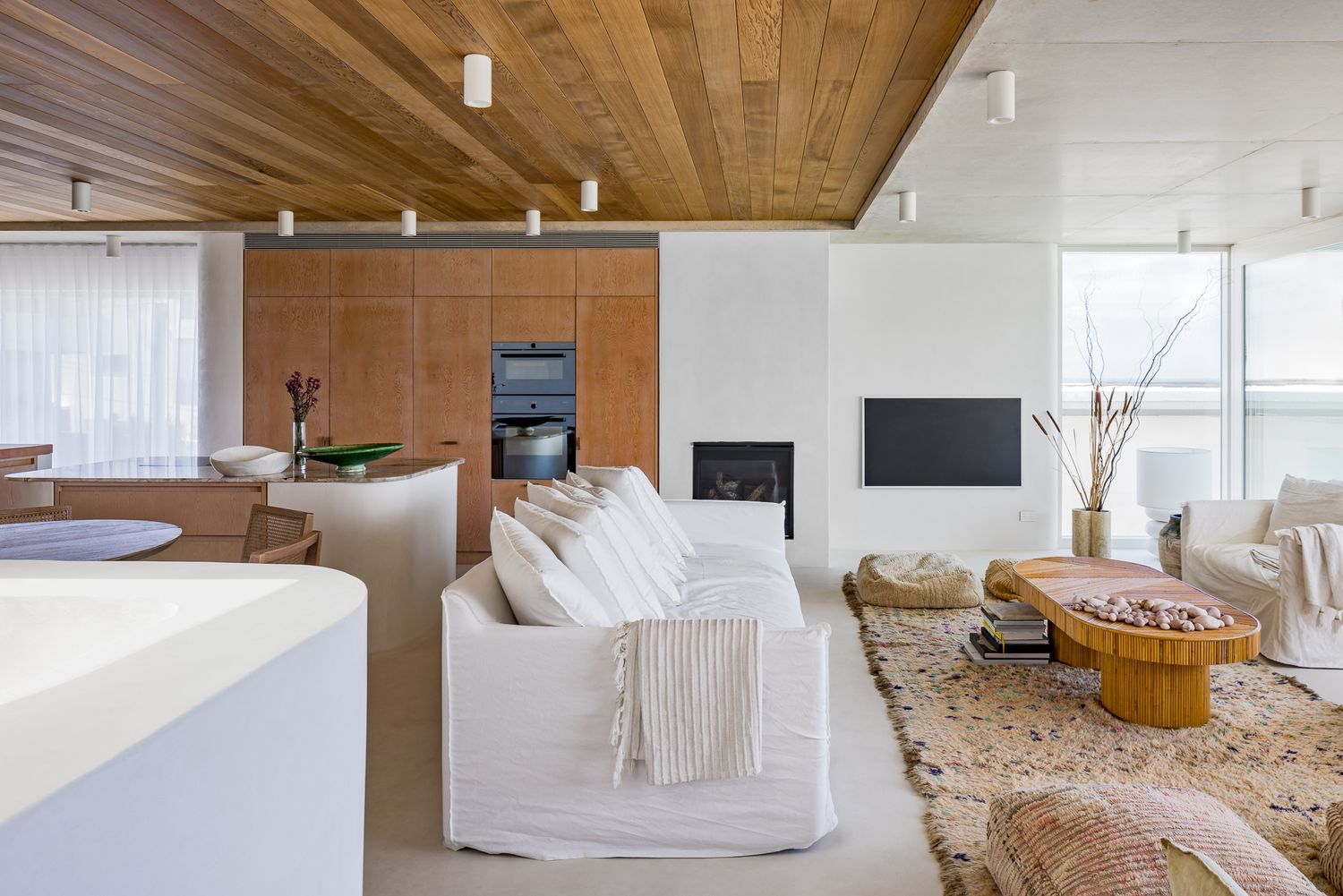 Open-plan living area with white linen sofas, timber ceiling, woven rug, fireplace and a kitchen with oak cabinetry beyond.