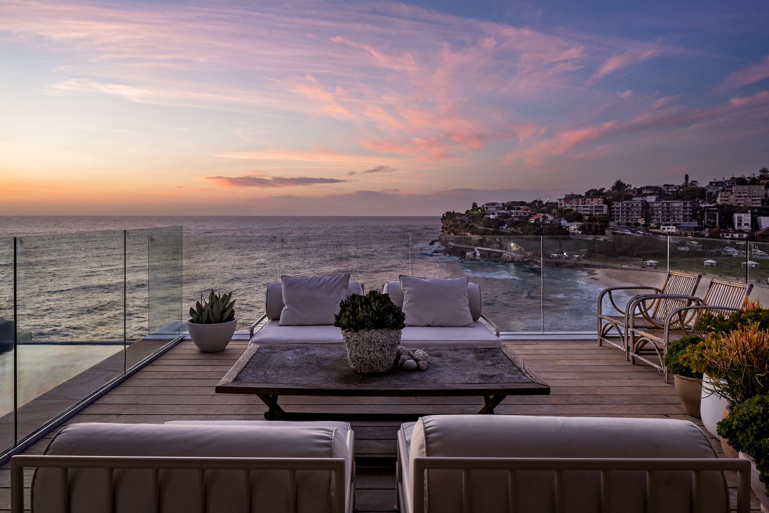 Rooftop seaside terrace with cushioned seating, a timber coffee table and glass railings overlooking a beach at sunset.