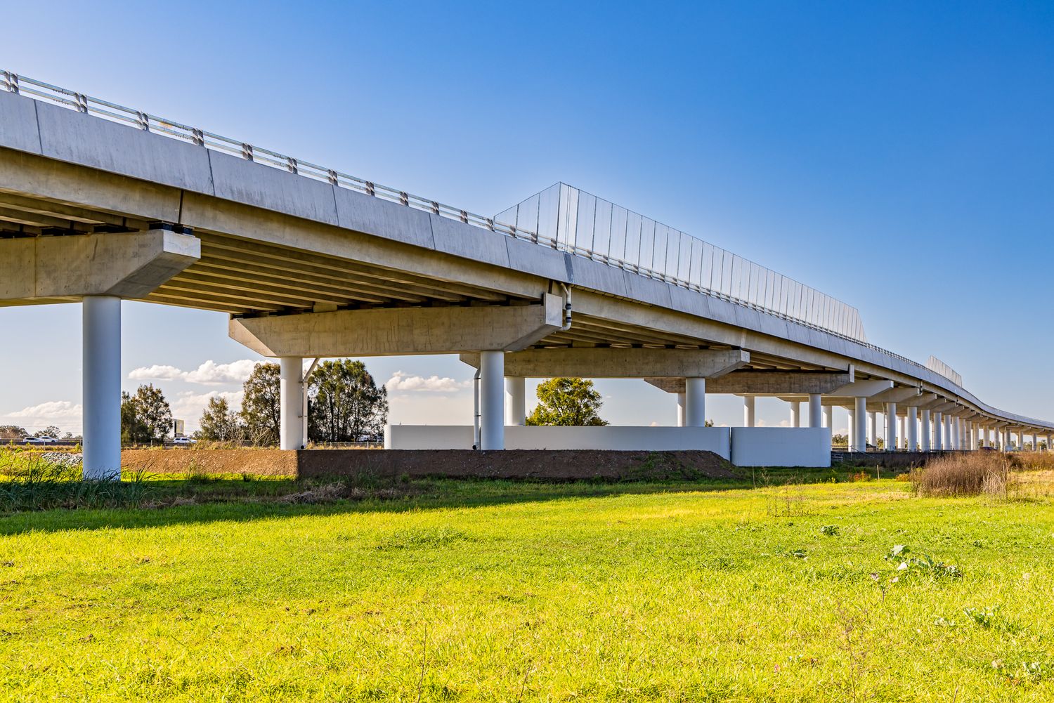Concrete overpass with tall support columns rising above a lush green field under a clear blue sky.