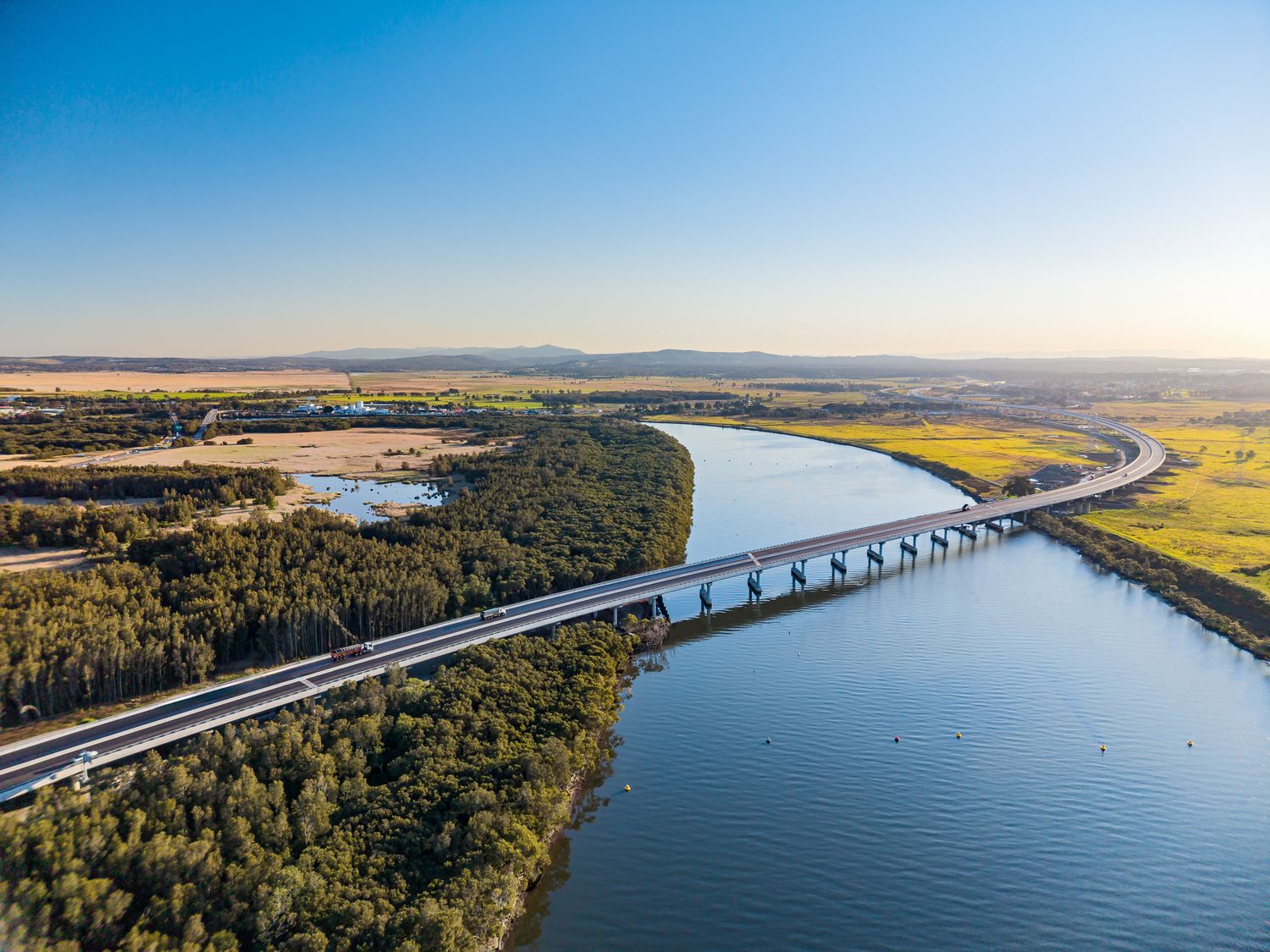 Aerial view of a highway bridge crossing a broad river, flanked by dense green trees and open fields in late afternoon light.