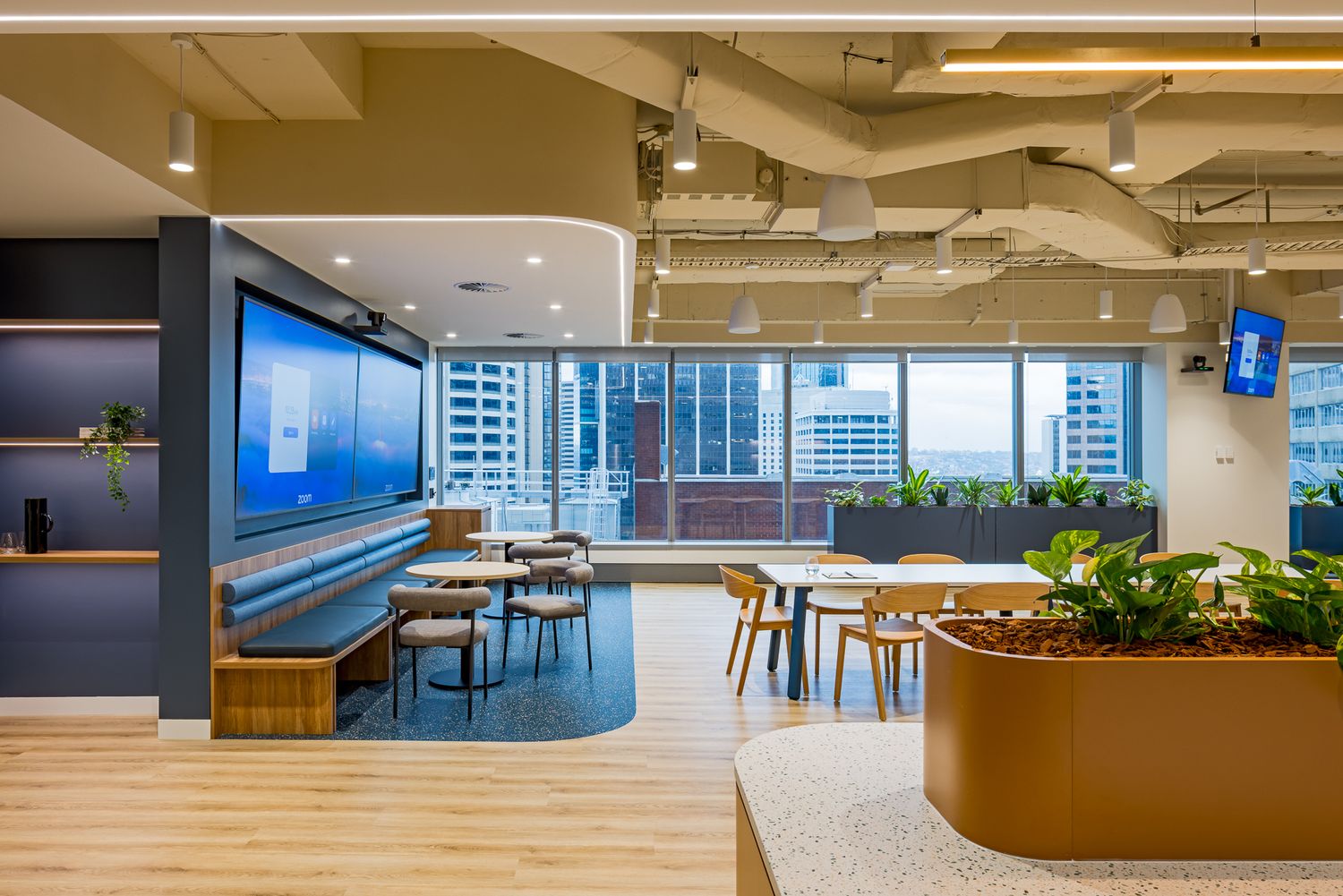 Modern office collaboration zone with banquette seating, wooden chairs, a planter box and wide windows overlooking city buildings.
