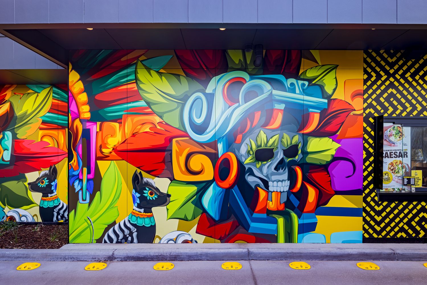 Close-up of a bright mural with a blue-headdressed skull, a jaguar and geometric patterns on a restaurant wall above yellow floor dots.