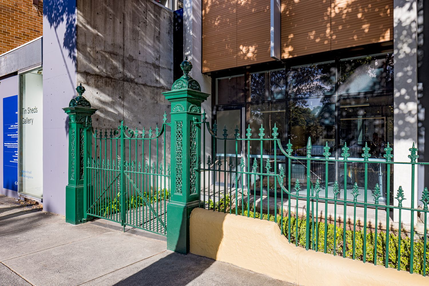 Green wrought iron fence and gate with decorative posts in front of a modern brick building on a sunny street.