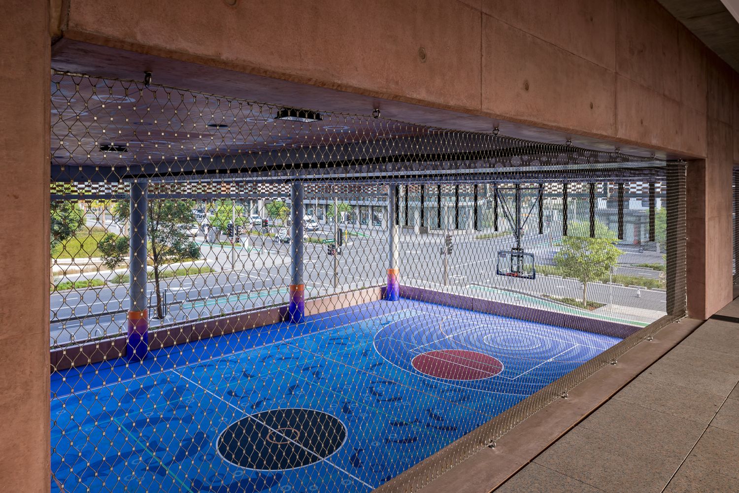 Rooftop basketball court with blue and red markings enclosed by mesh fencing, overlooking a tree-lined street below.