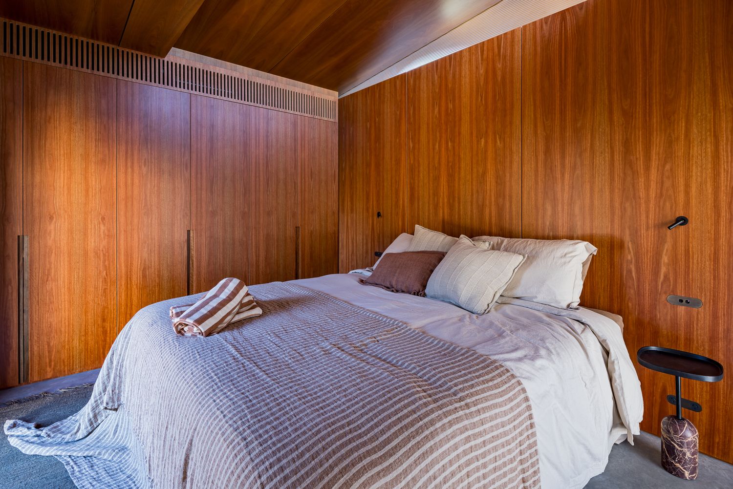 Cozy bedroom with warm timber-paneled walls and ceiling, a made bed with striped throw and pillows, and a small side table.