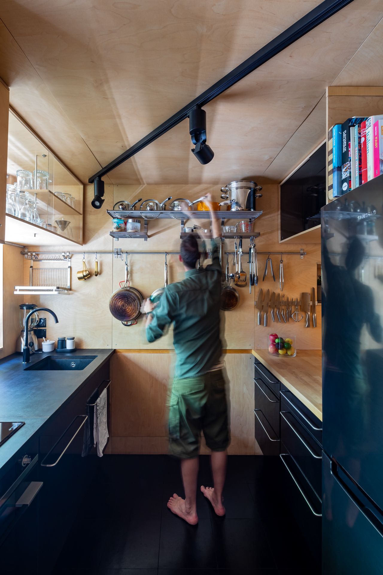 Person reaching for hanging pots in a compact plywood kitchen with black cabinetry, track lighting and a steel fridge.