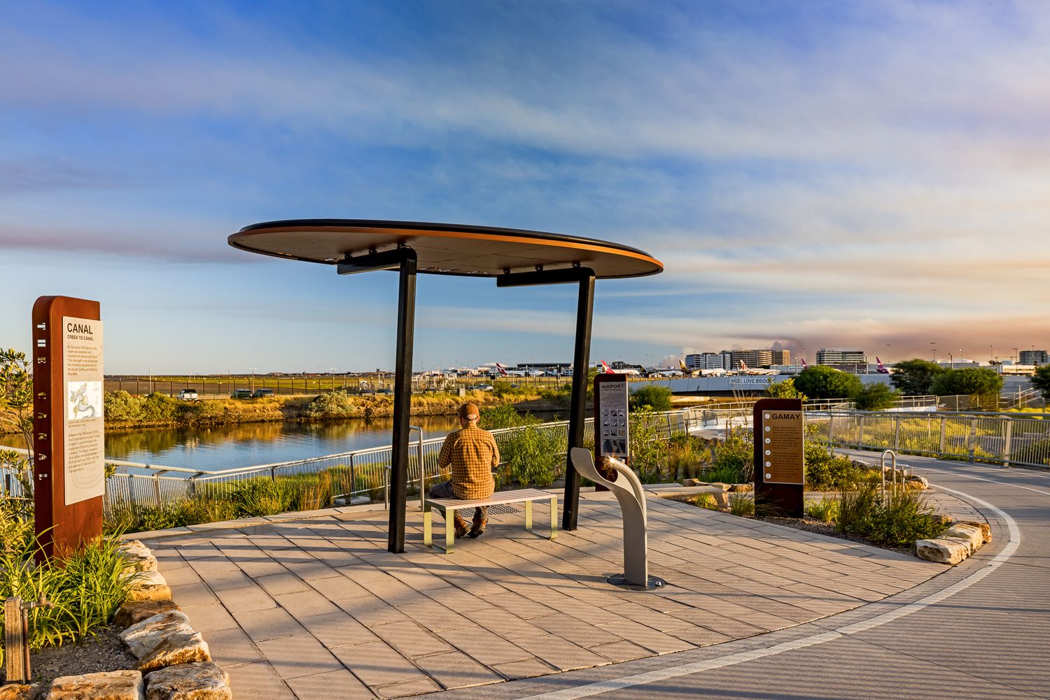 Person sits under a round shade canopy on a paved waterfront path with interpretive signs, stone edging and city views at dusk.
