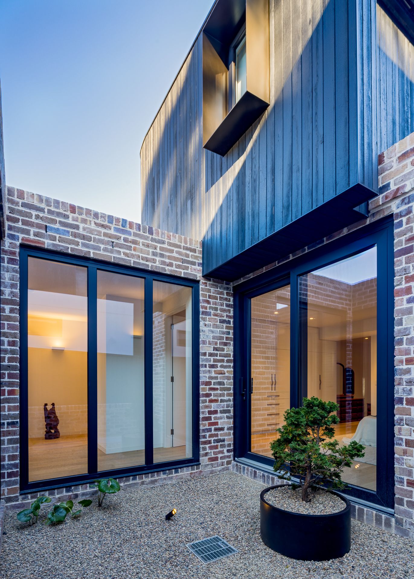 Brick courtyard with black-framed glass doors, dark timber cladding above and a pine bonsai in a round black planter on gravel.