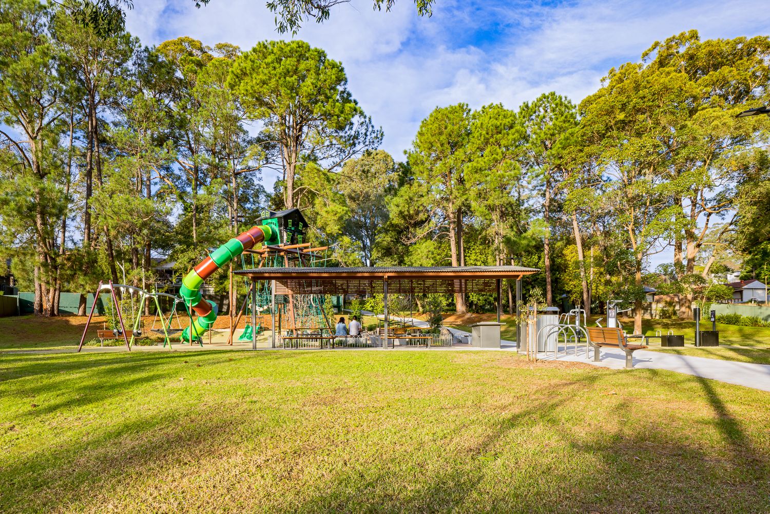 Green and orange spiral slide on a treehouse-style play tower beside a covered picnic shelter on a sunny grassy park lawn.