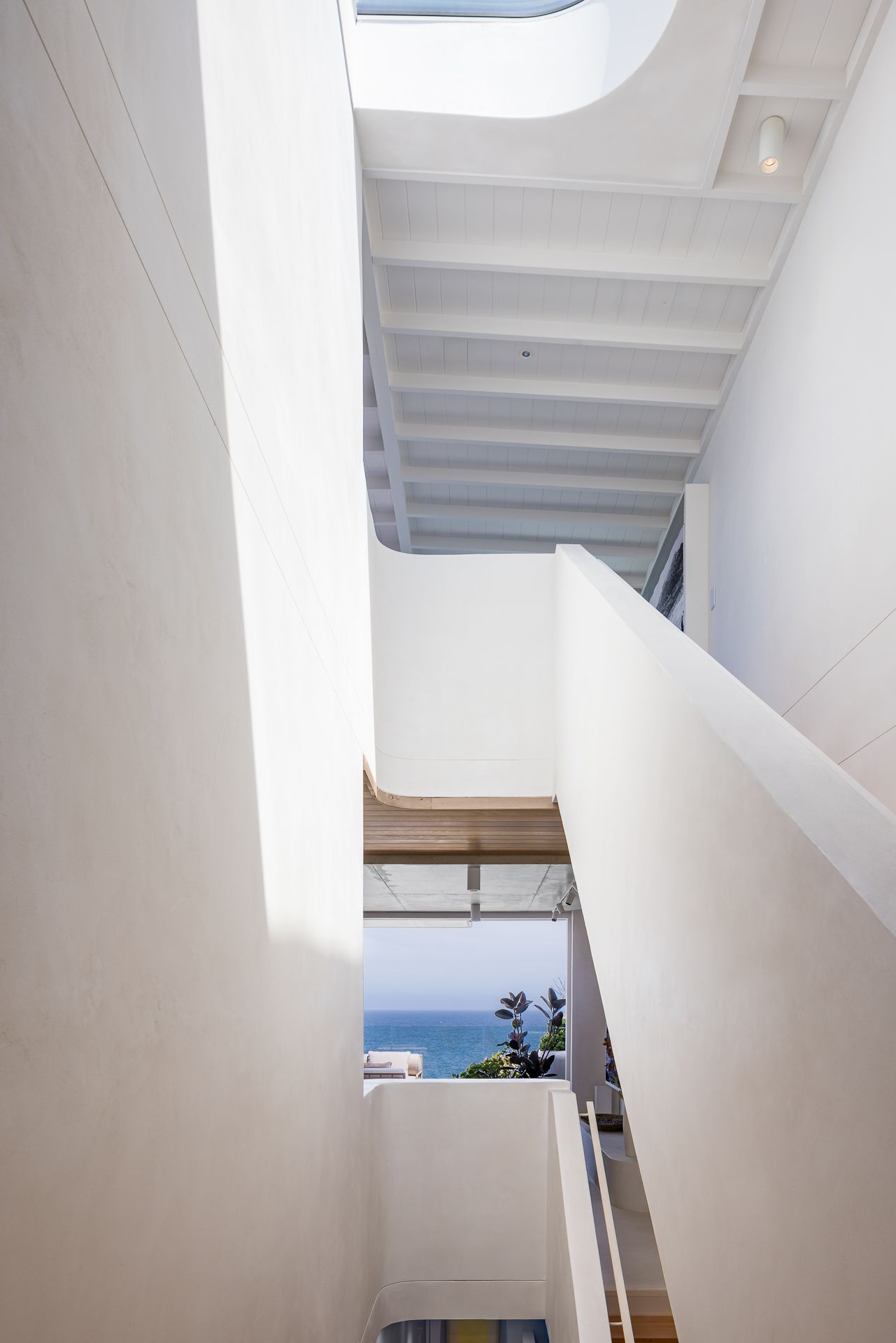 Bright white architectural stairwell with sculpted curved walls and a skylight, framing a glimpse of the blue ocean below.