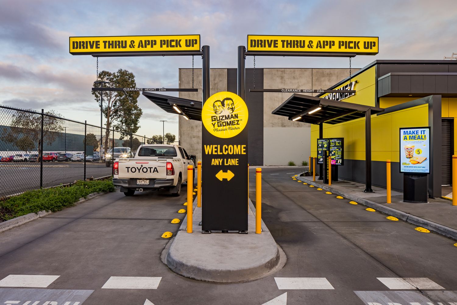 Guzman y Gomez drive-thru lane with Drive Thru App Pick Up signs, a welcome sign and a white pickup truck waiting to order.