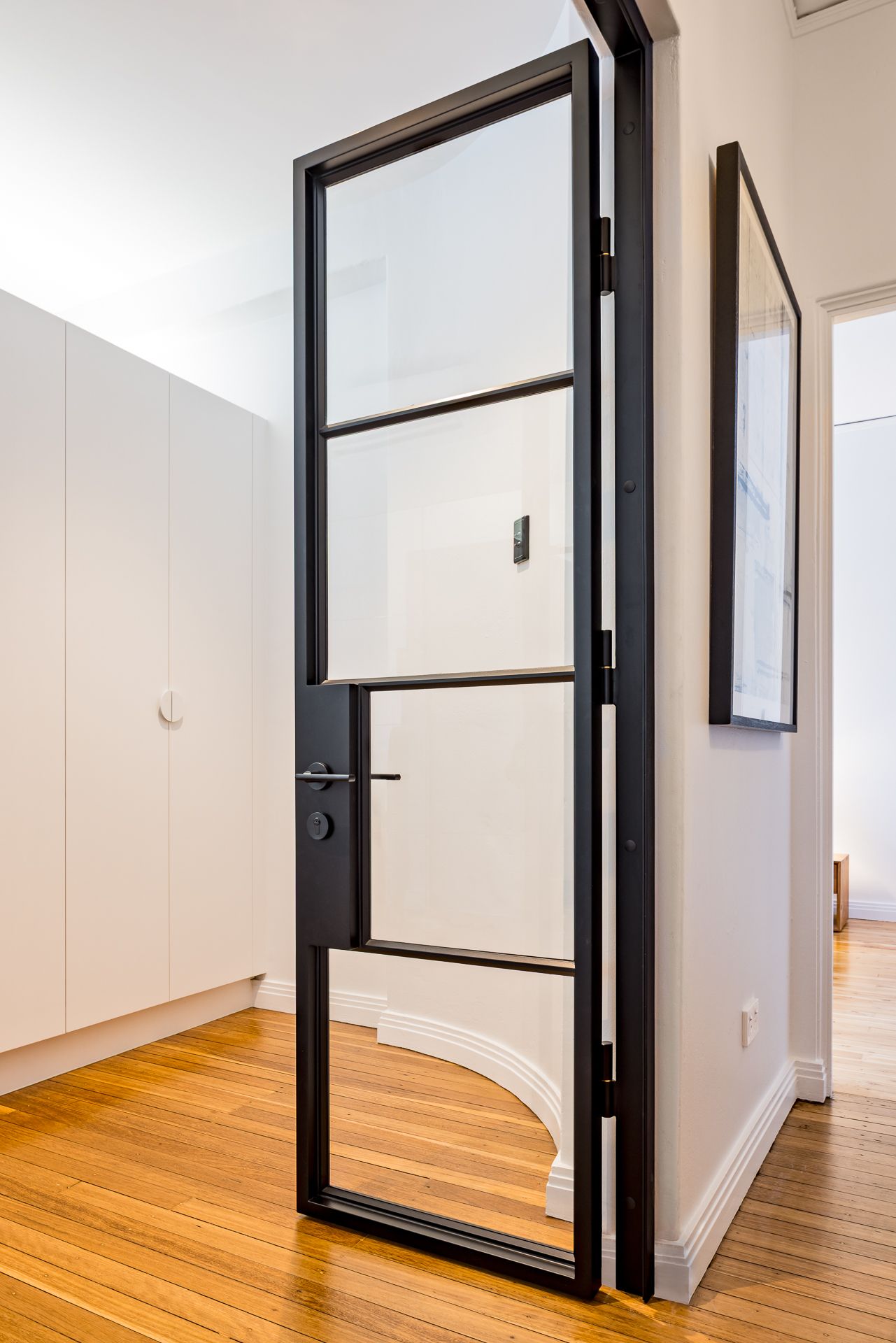 Black steel-framed glass door in a white hallway with timber floors, built-in white cupboards and framed artwork.