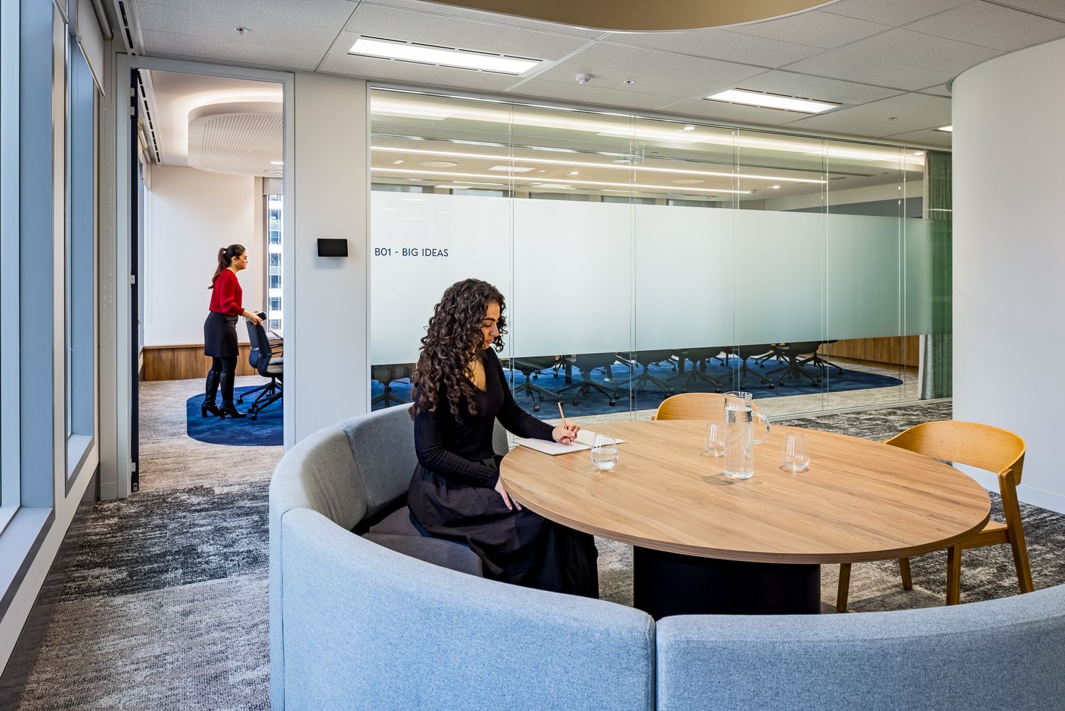 Woman in black sits at a round wooden table in a curved gray booth inside a bright office meeting nook with glass walls.